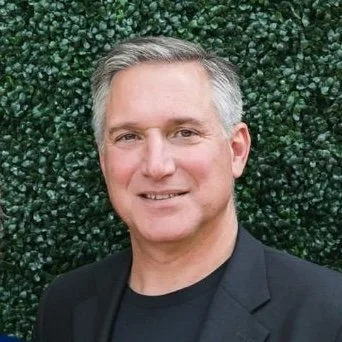 A middle-aged man with short gray hair, wearing a black blazer and black t-shirt, smiling in front of a green leafy background.
