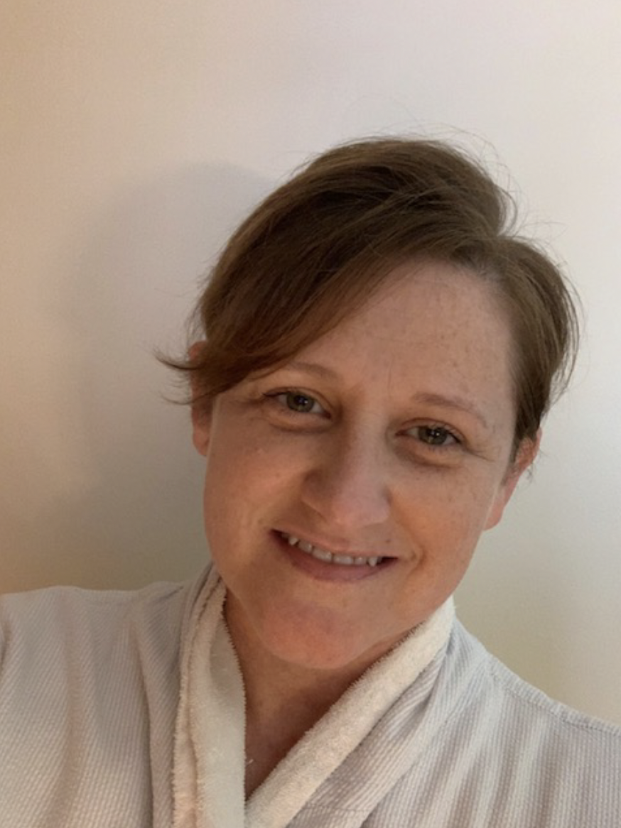 A person with short brown hair smiling in front of a plain light-colored wall, wearing a light-colored robe.