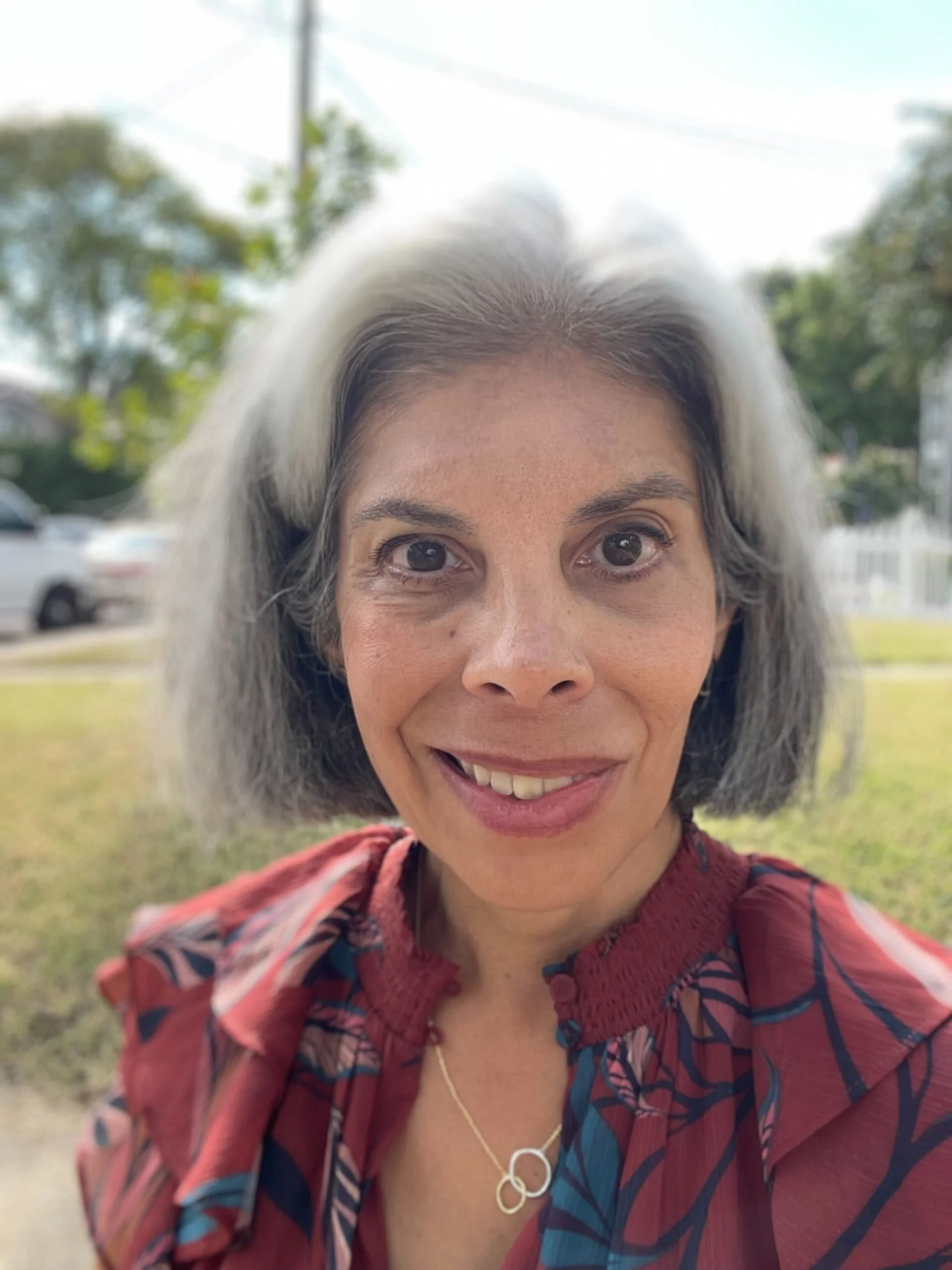 A woman with gray hair and brown eyes smiling outdoors, wearing a red patterned blouse and a gold necklace, with a blurred background of trees, cars, and a fence.