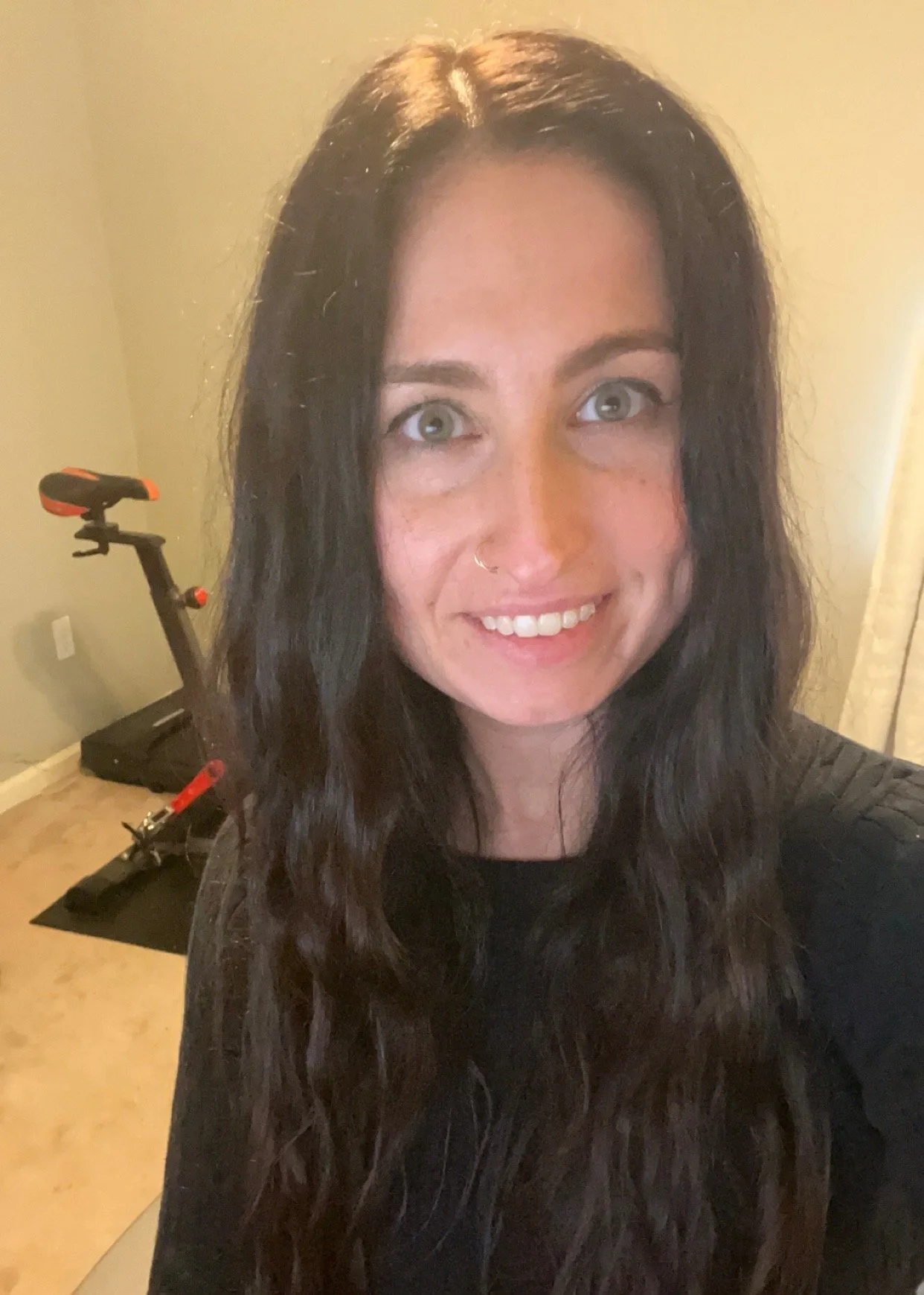 A woman with long dark wavy hair, light skin, and green eyes smiling at the camera, in a room with a beige wall and a treadmill in the background.