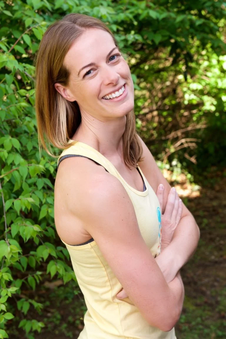 A woman with light brown hair smiling outdoors, standing in front of green leafy bushes, wearing a yellow sleeveless top.