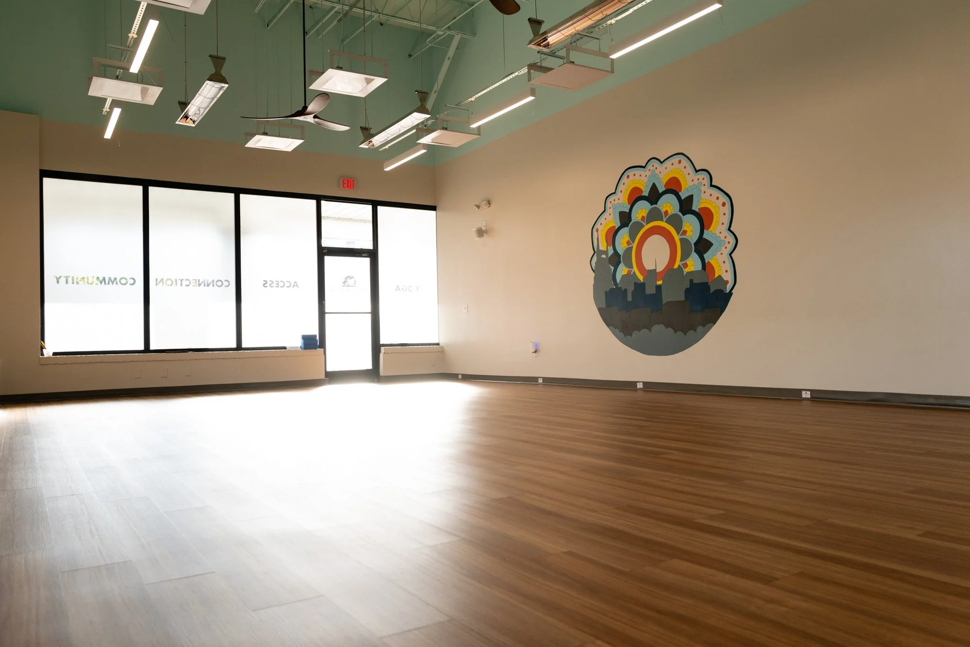 Empty room with hardwood floors, large windows, colorful mural of a cityscape with a sunrise and decorative elements on the wall, ceiling fans, and modern lighting.