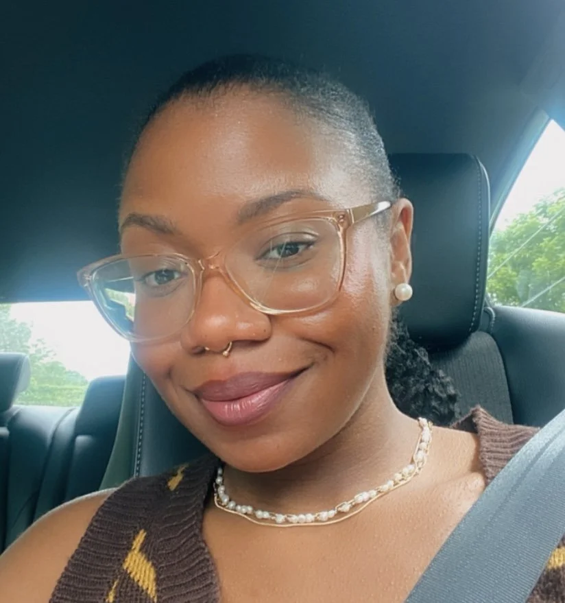 A woman with glasses and pearl earrings sitting in a car, smiling at the camera, with greenery visible outside the window.