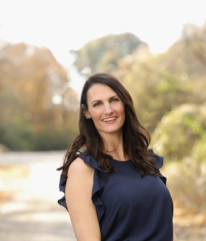 Woman with long dark hair smiling outdoors during daytime, wearing a navy sleeveless top with ruffled details, standing in front of a blurred background of trees with autumn foliage.
