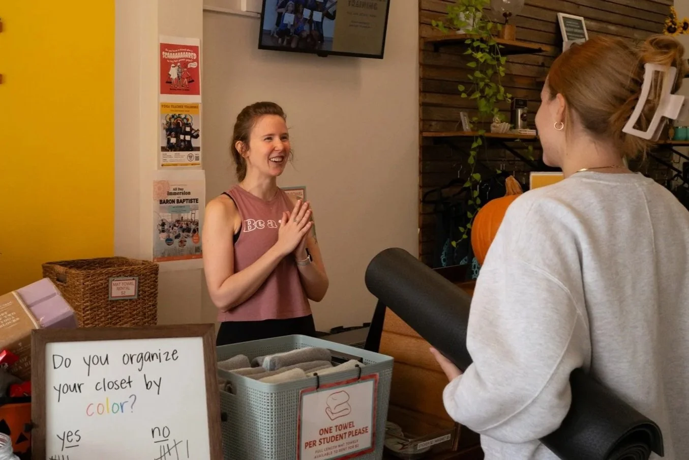 A woman at reception desk smiling and talking to a female visitor who is holding a yoga mat at a yoga studio.