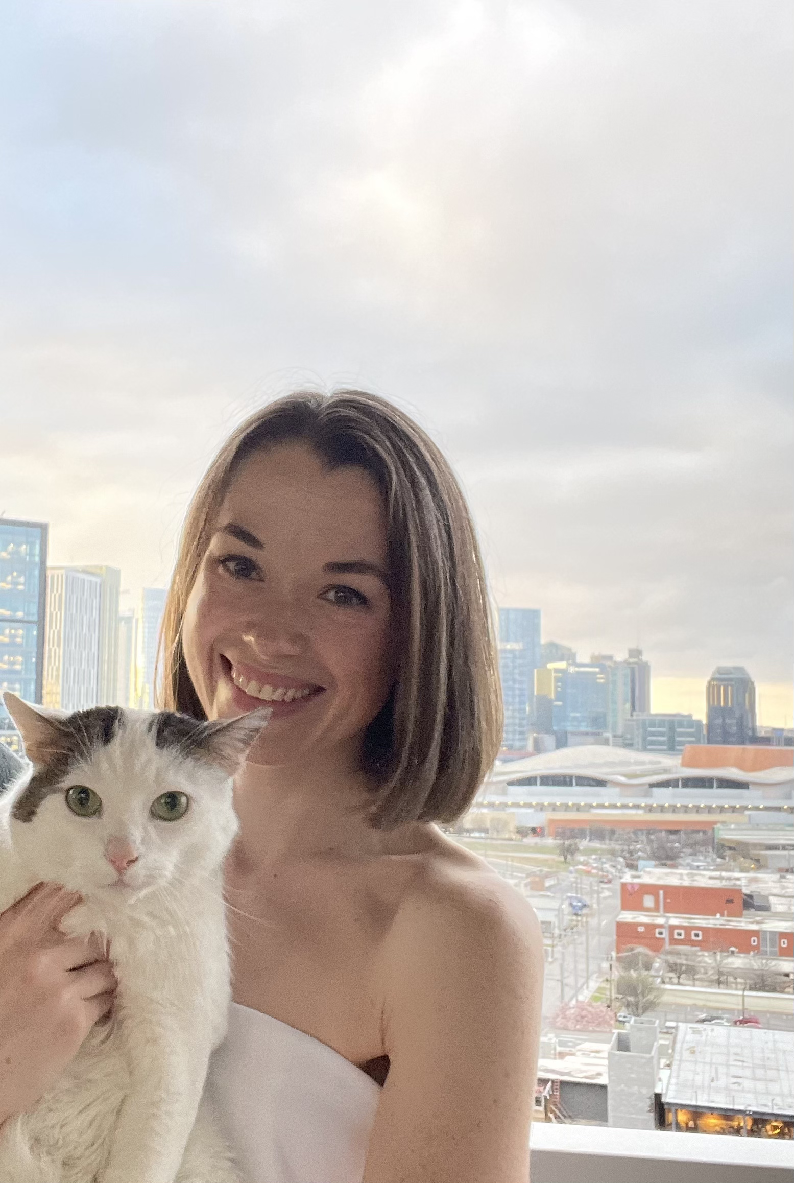 A smiling woman is holding a white and gray cat with green eyes in front of a city skyline window.