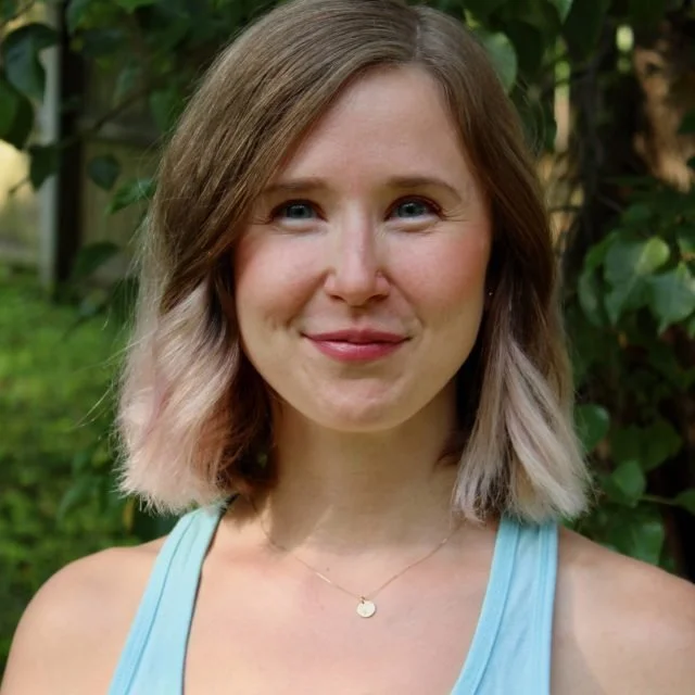 Close-up of a young woman with shoulder-length light brown hair with blonde tips, smiling slightly, wearing a light blue tank top and a delicate necklace, outdoors with green foliage in the background.