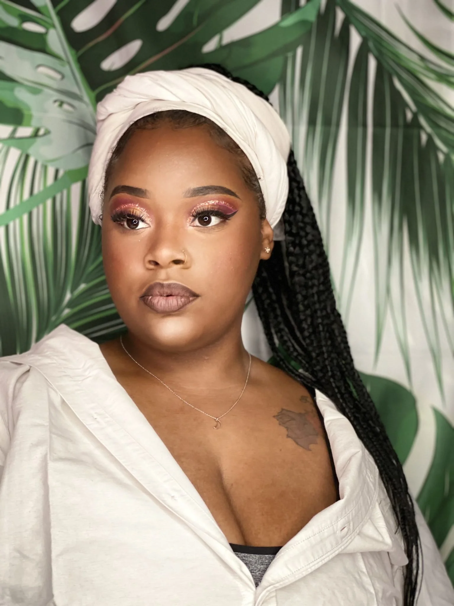 A woman with dark hair in braids, wearing a white headwrap and a white jacket, poses in front of a green tropical leaf background.