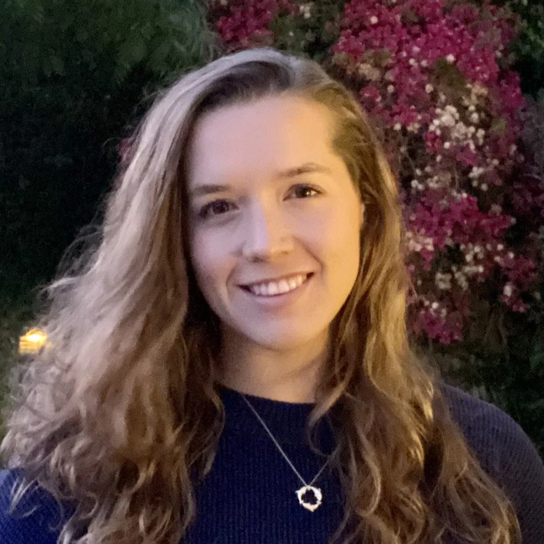 A young woman with long, wavy hair smiling outdoors at dusk, standing in front of a bush with pink and white flowers.