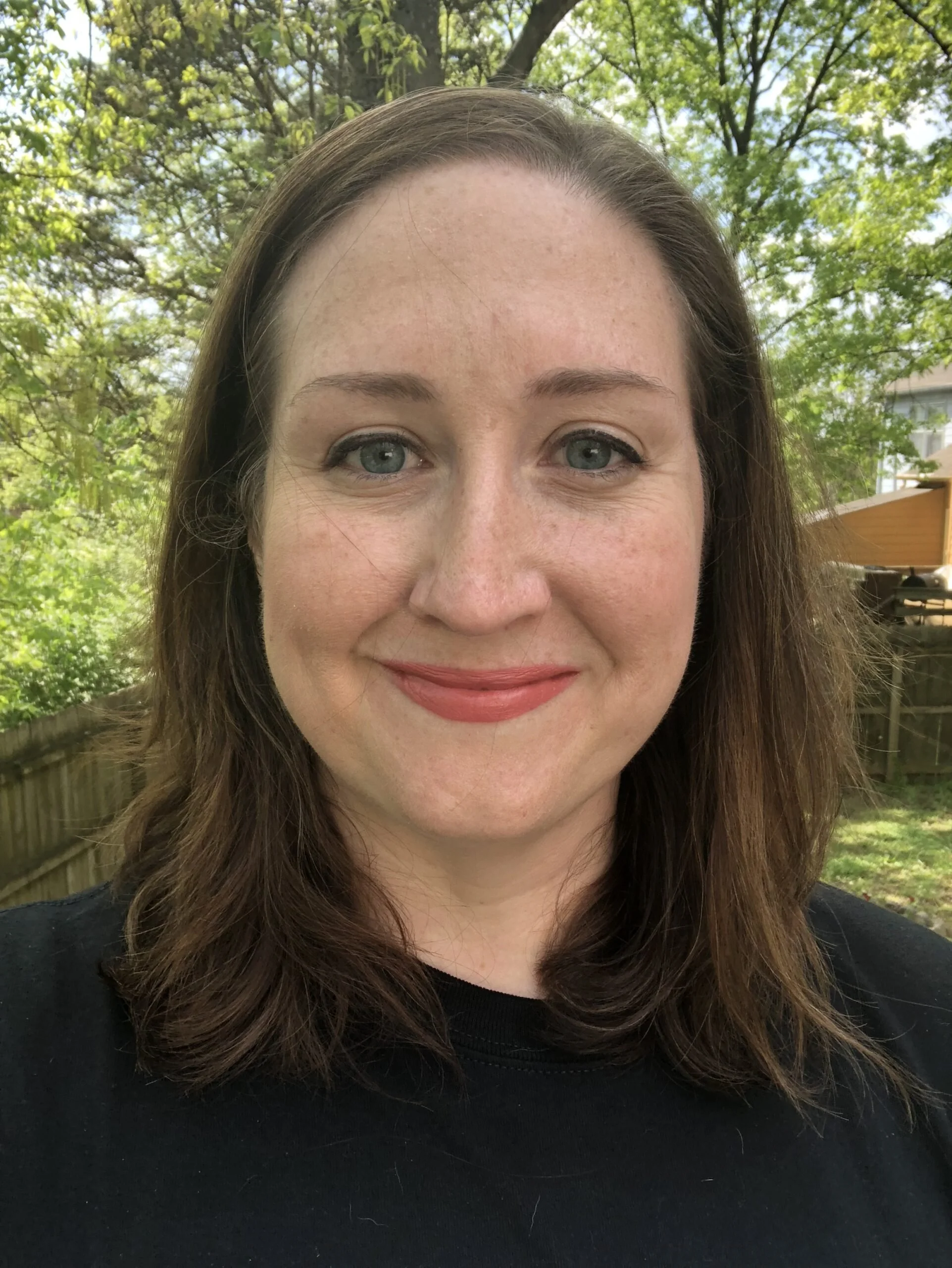 A woman with shoulder-length brown hair and blue eyes smiling outdoors with trees and a wooden fence in the background.