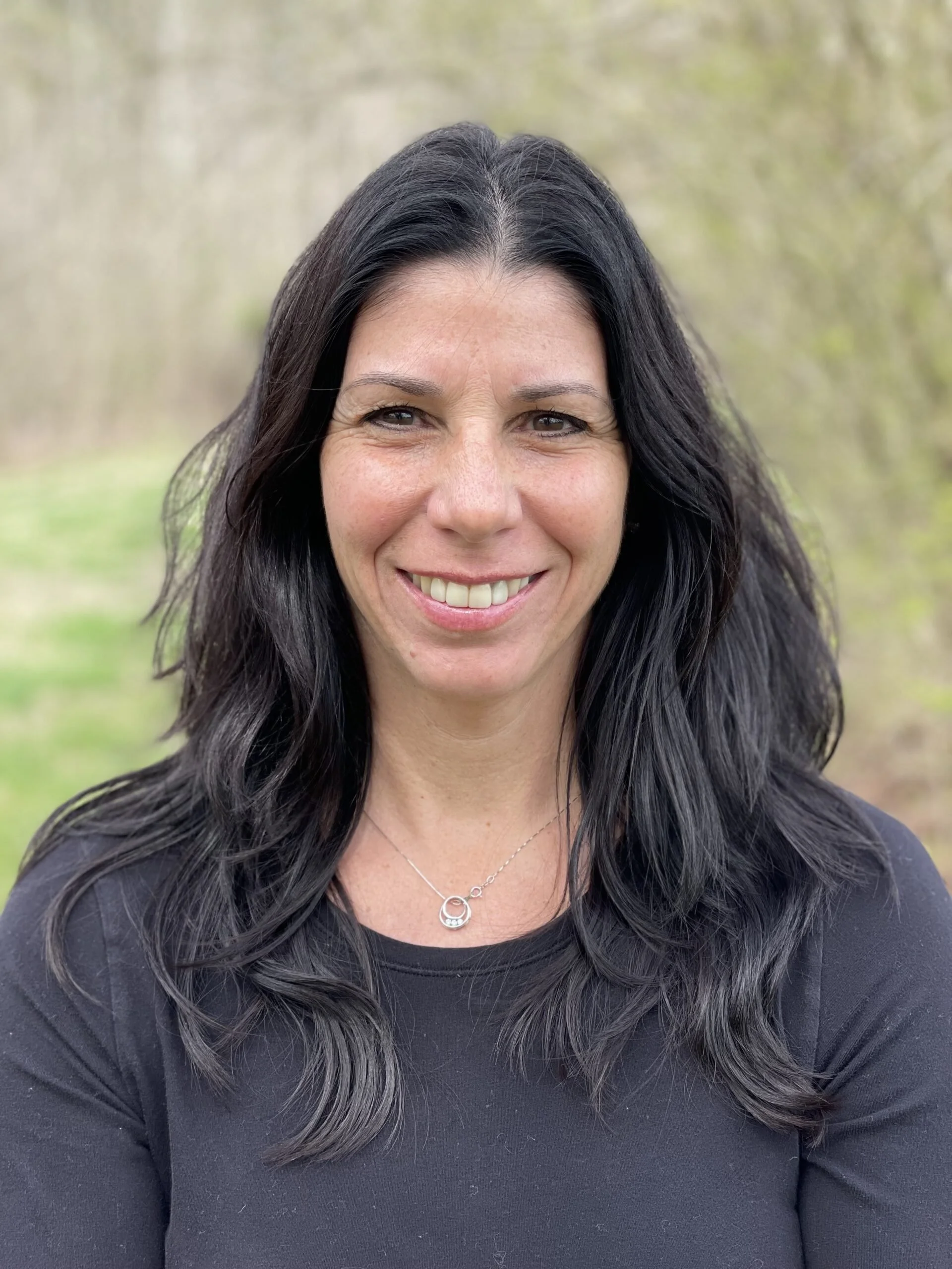 A woman with long black hair, wearing a black top and a silver necklace, smiling outdoors with a blurred background of trees and grass.