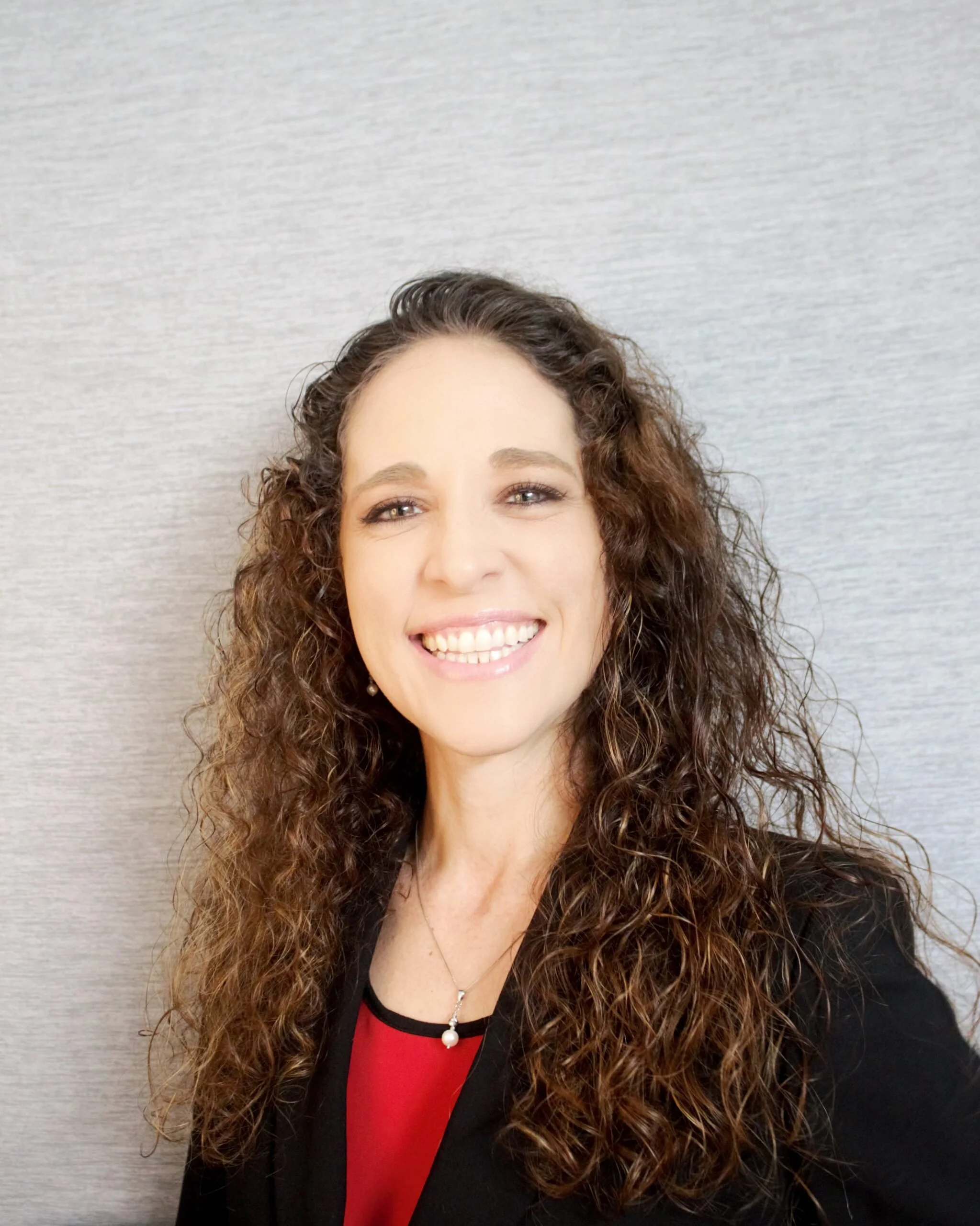 A woman with long, curly brown hair smiling against a light gray background, wearing a black blazer, red blouse, and pearl jewelry.