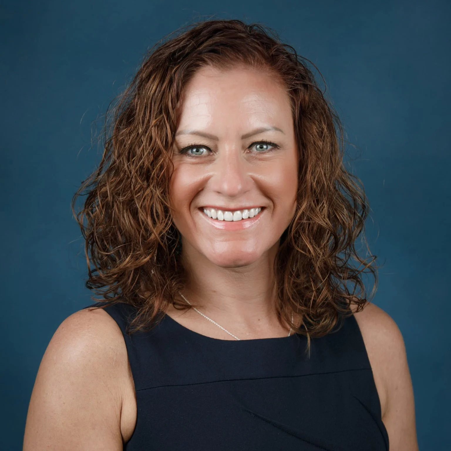 A woman with curly brown hair smiling at the camera, wearing a sleeveless black top against a dark blue background.
