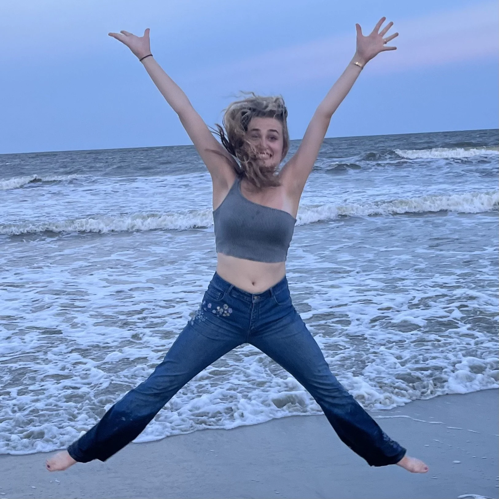 A young woman jumping with arms and legs spread wide on a beach, smiling, with ocean waves in the background during what appears to be dusk or twilight.