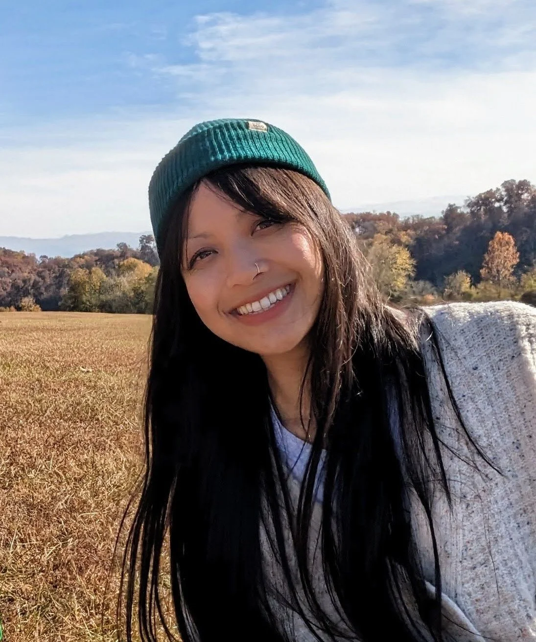 A smiling woman with a nose ring, long black hair, wearing a green beanie and a gray sweater, outdoors in a field with trees and hills in the background.
