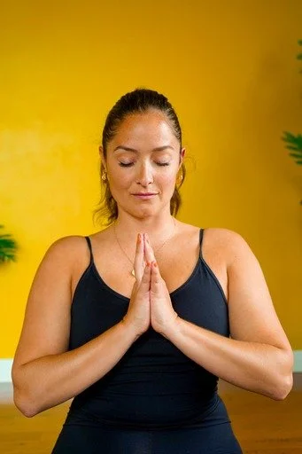 A woman practicing yoga with her hands in a prayer position, eyes closed, against a yellow background.