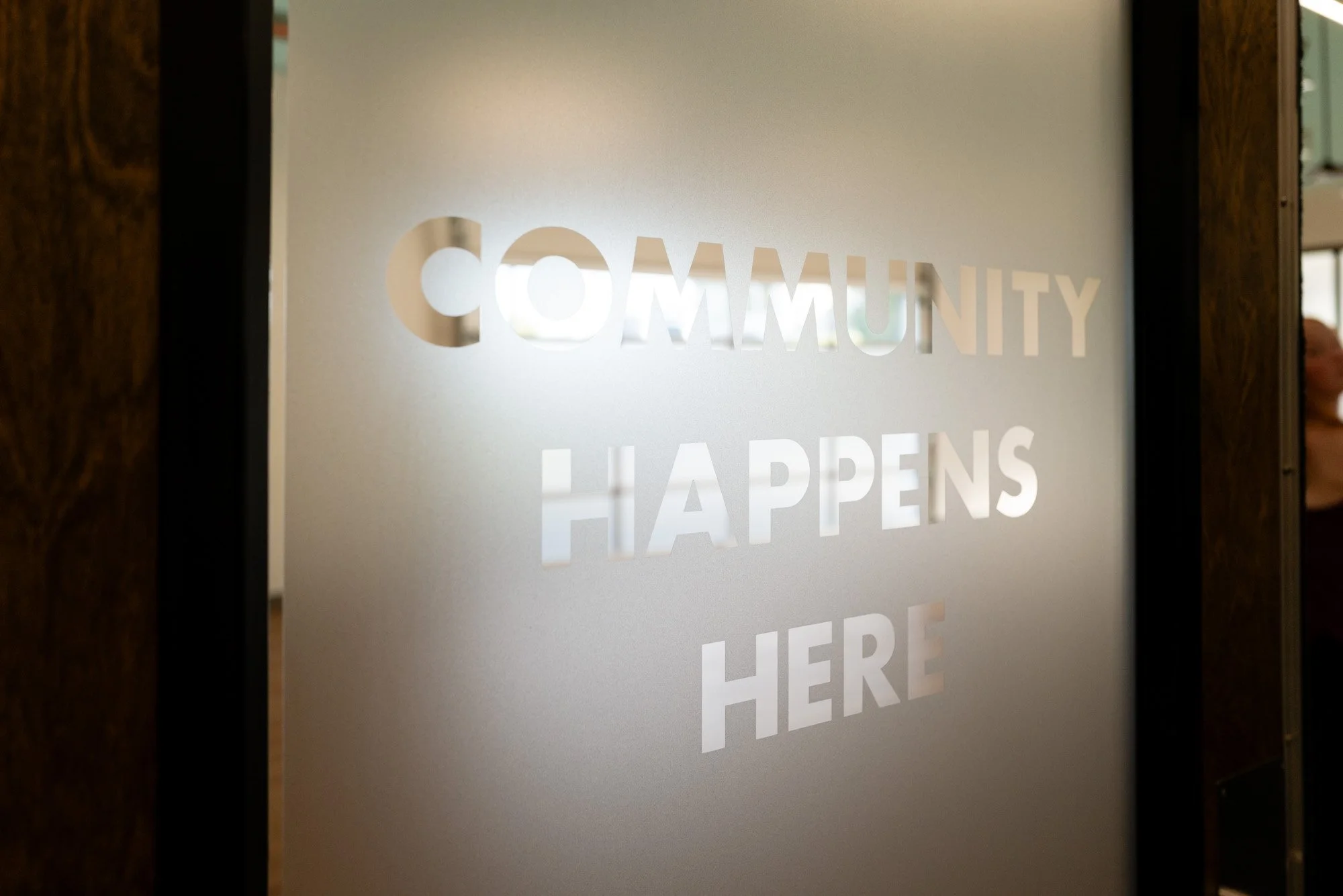 Frosted glass door with the words 'Community Happens Here' cut out, partially revealing a room behind.