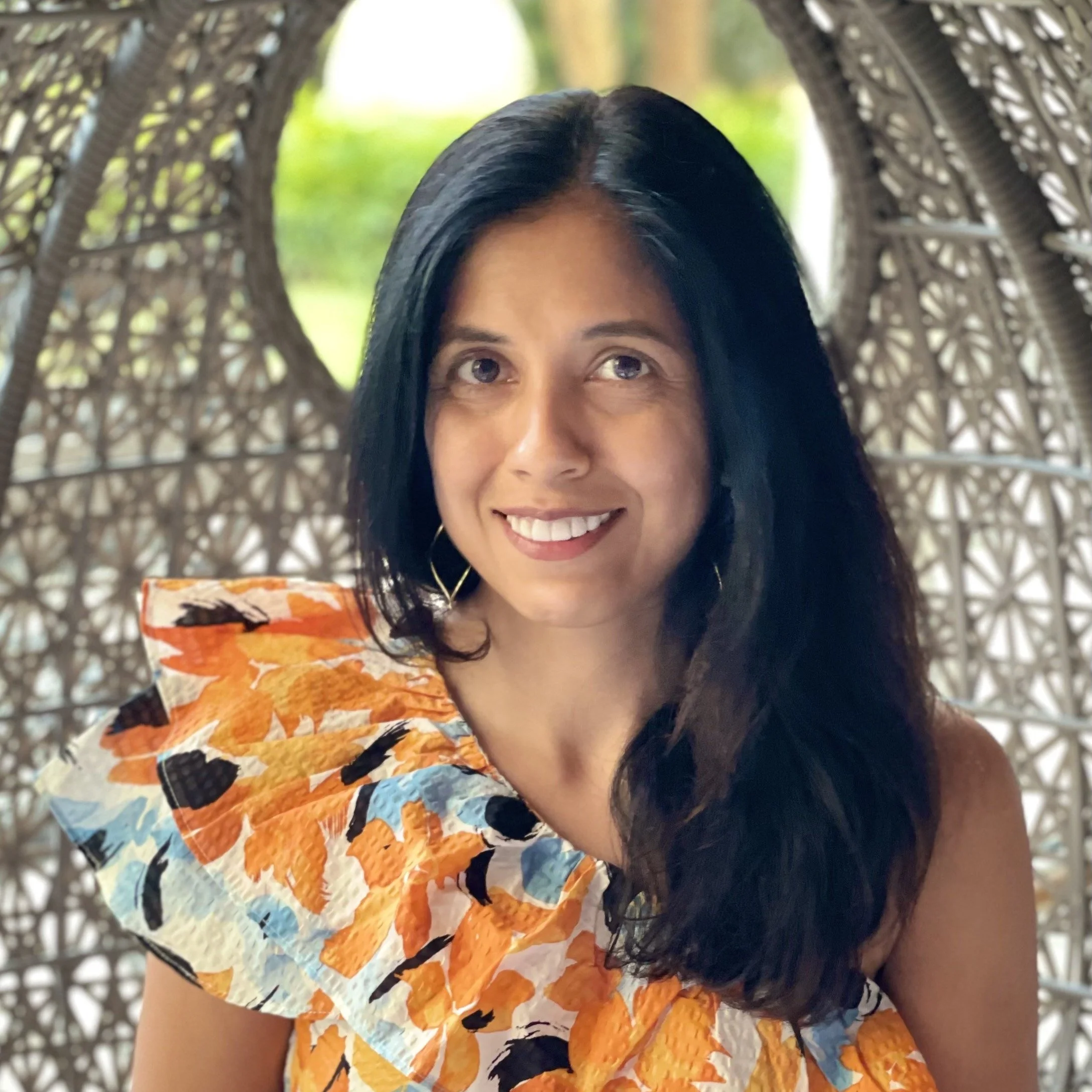 A woman with long black hair smiling, wearing an orange dress with ruffled sleeves with a floral pattern, sitting in front of a decorative woven background with green outdoor scenery in the distance.