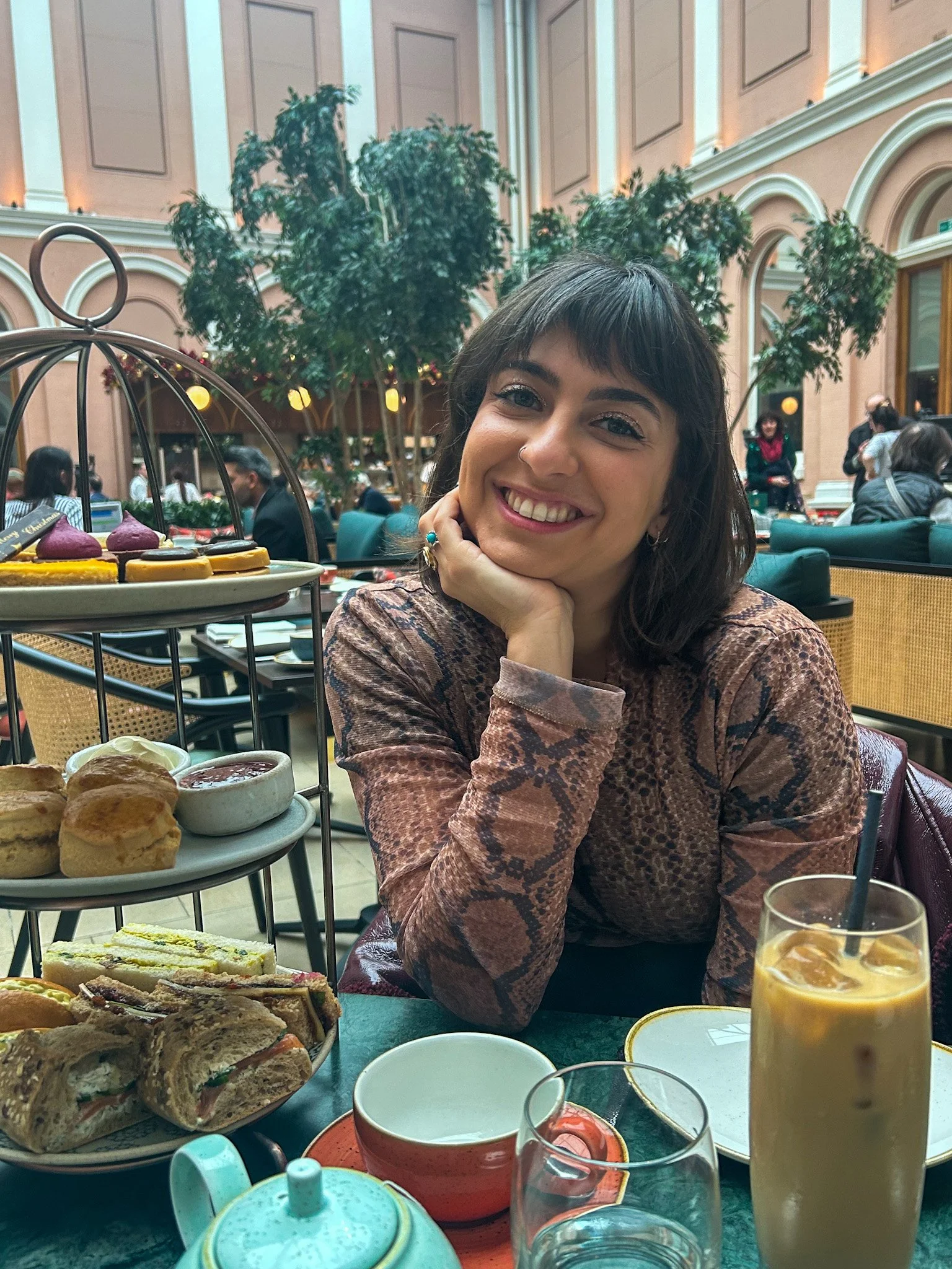 A woman with short dark hair and a snake print top sitting at a table in a fancy indoor restaurant, smiling with her head resting on her hand. The table has a tiered stand of scones, sandwiches, and desserts, along with a glass of iced coffee and some cups.