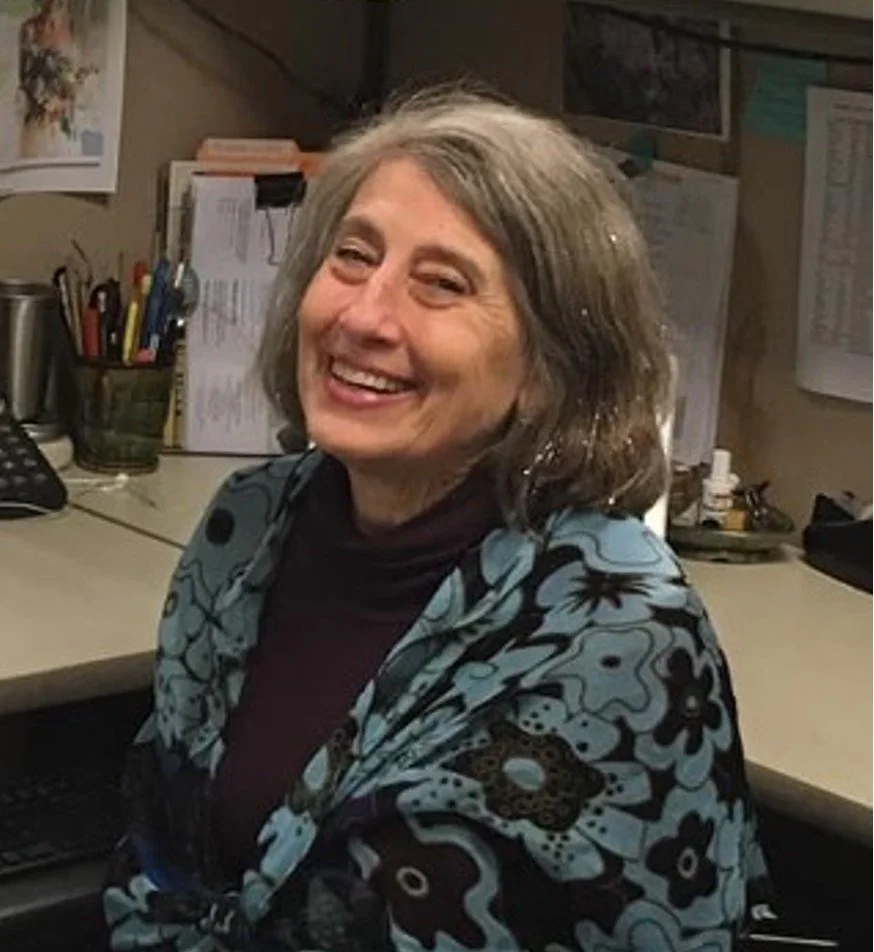 A smiling woman with gray hair sitting at an office desk with various papers, office supplies, and a telephone behind her.