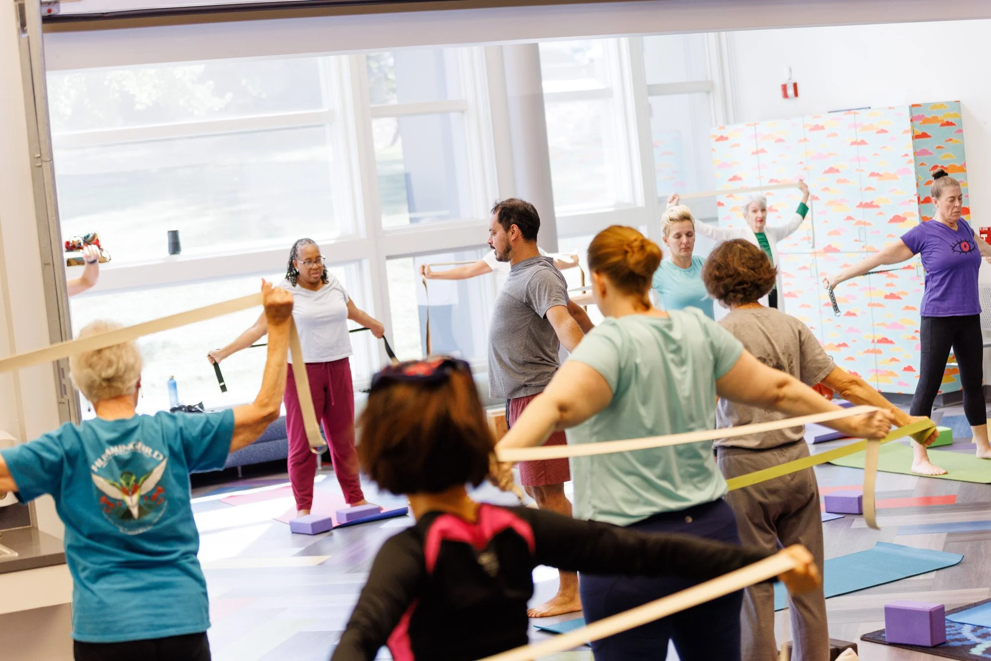 A group of people participating in a group exercise or yoga class using resistance bands in a well-lit studio.