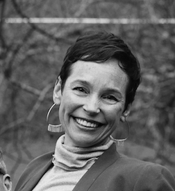 Black and white portrait of a woman smiling outdoors, wearing hoop earrings and a turtleneck.