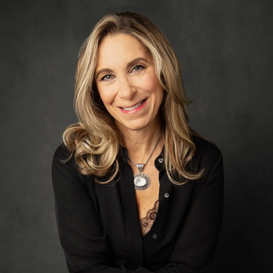 A woman with long blonde hair, smiling, wearing a black shirt and a silver pendant necklace, posing against a dark background.