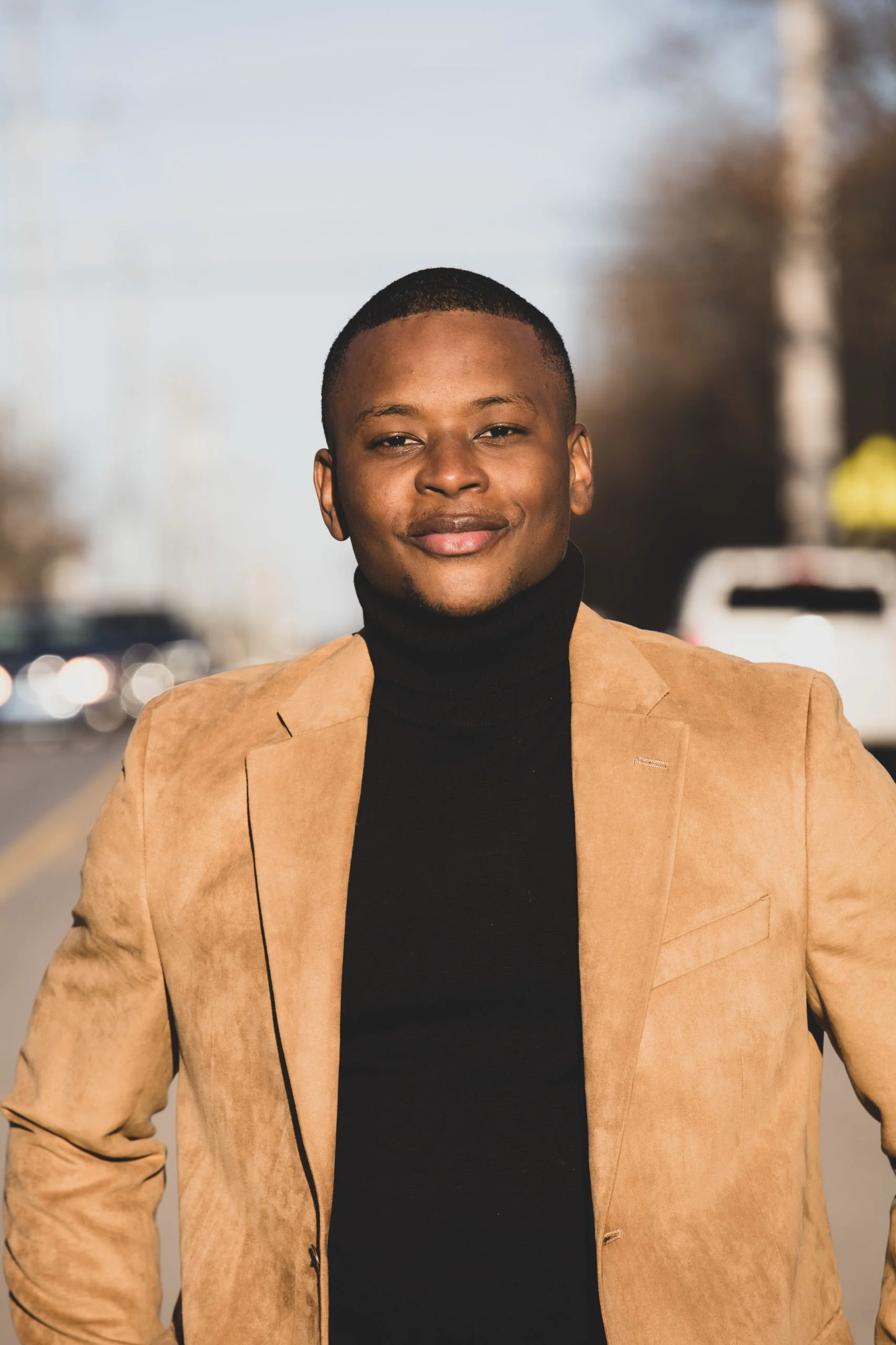 A young man, smiling, wearing a tan blazer and black turtleneck, standing outdoors on a street with cars and trees in the background.
