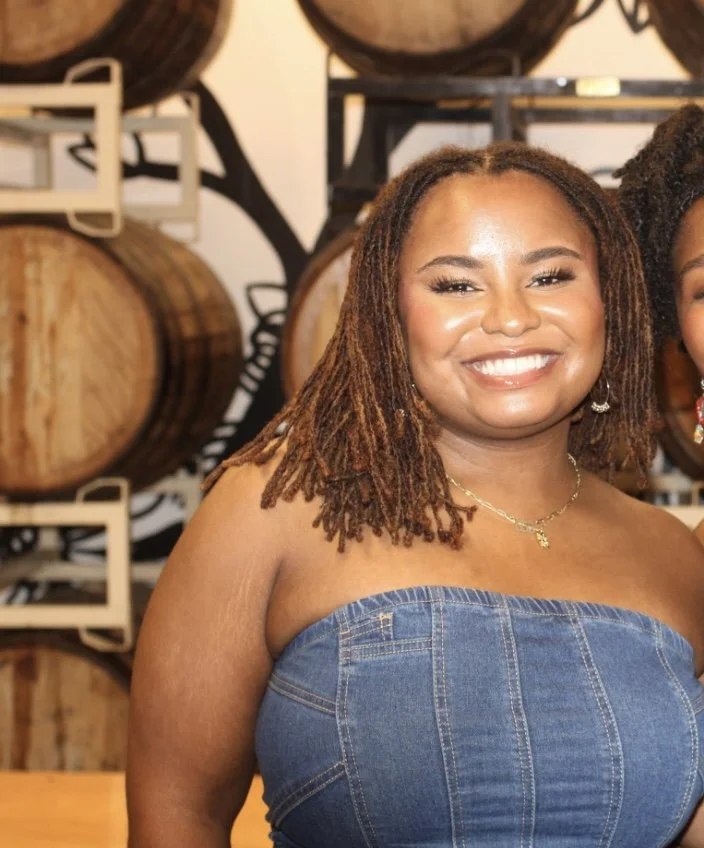 A smiling woman with shoulder-length dreadlocks wearing a strapless denim dress and gold jewelry standing in front of stacked barrels at a wine or whiskey cellar.