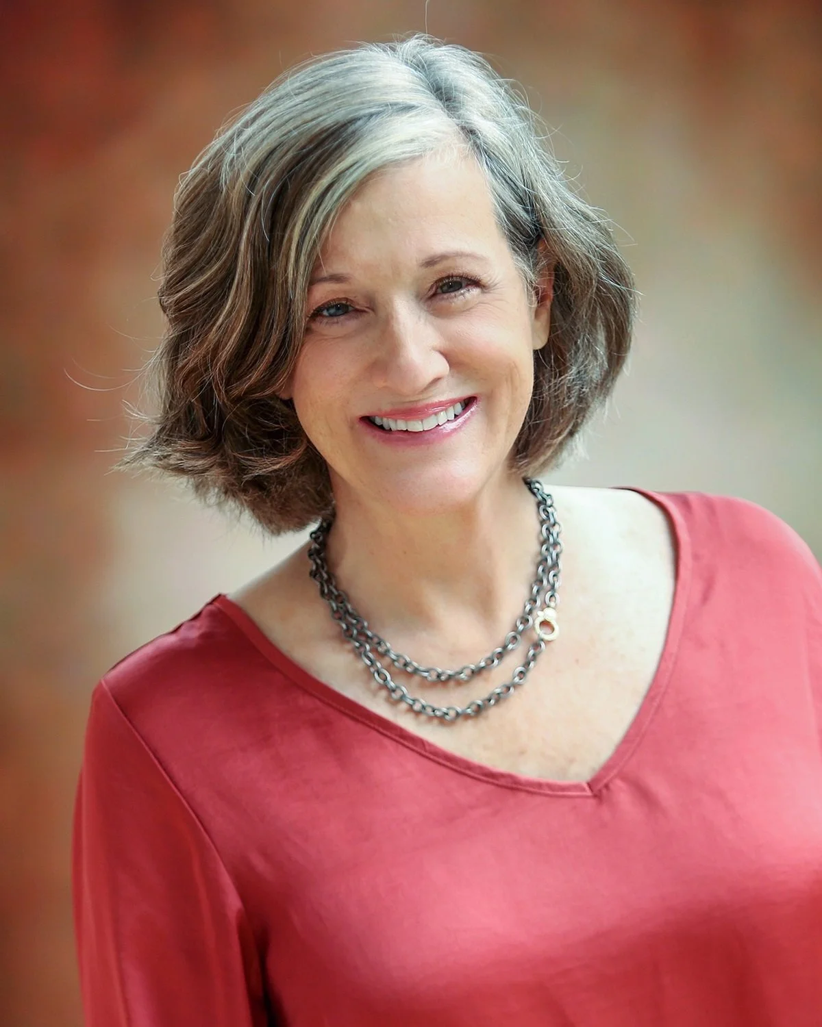 A middle-aged woman with shoulder-length gray hair, smiling, wearing a red top and layered silver necklaces.
