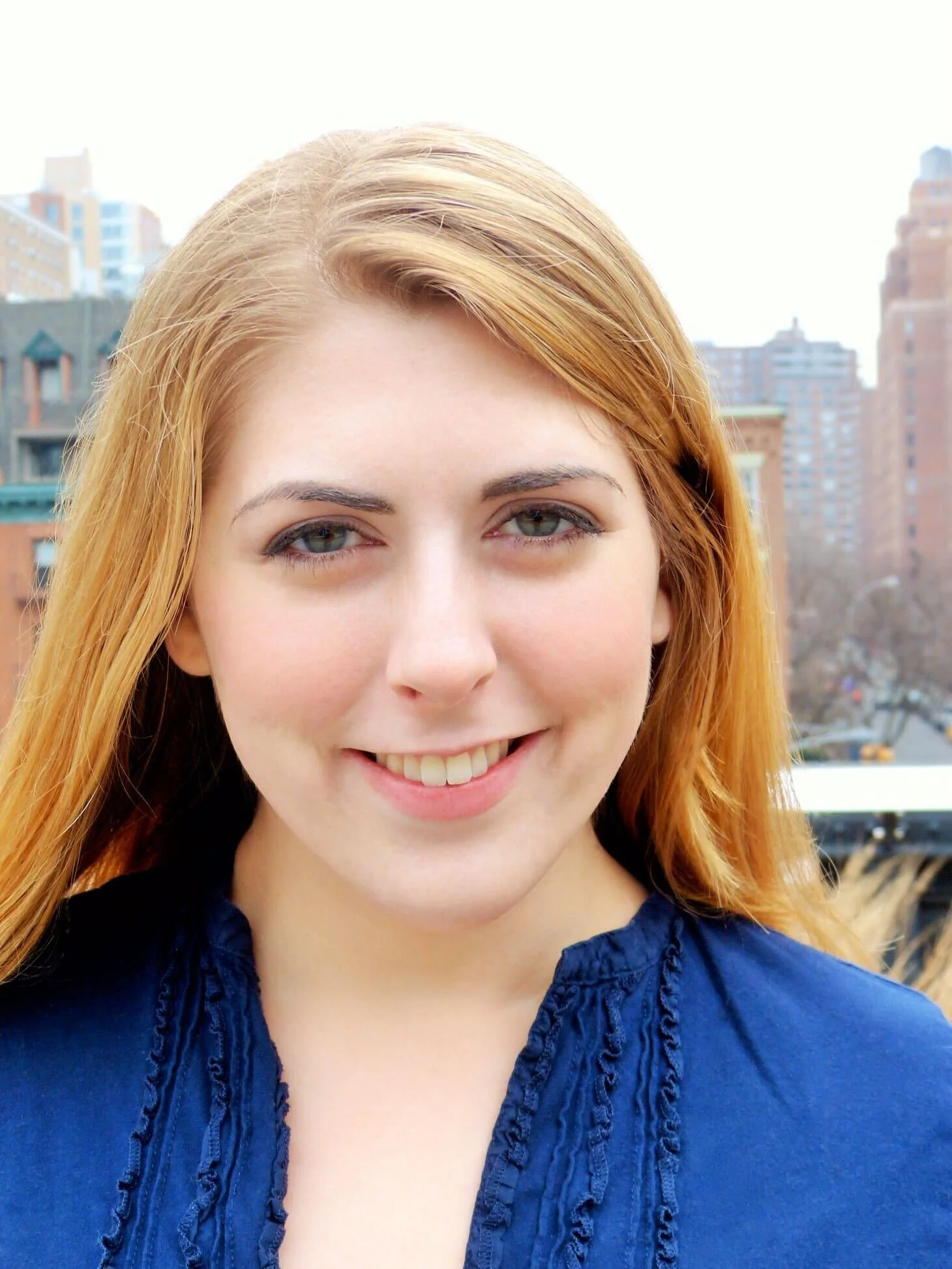 Close-up of a smiling woman with red hair outdoors with city buildings in the background.