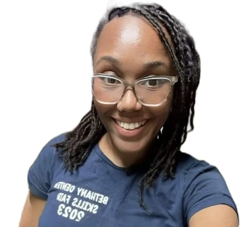 A woman with dark, twisted dreadlocks, glasses, and a big smile, wearing a blue t-shirt with white text, against a plain white background.