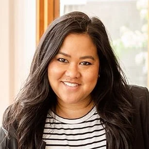 Portrait of a woman with long black hair smiling inside a room with wooden window frames.