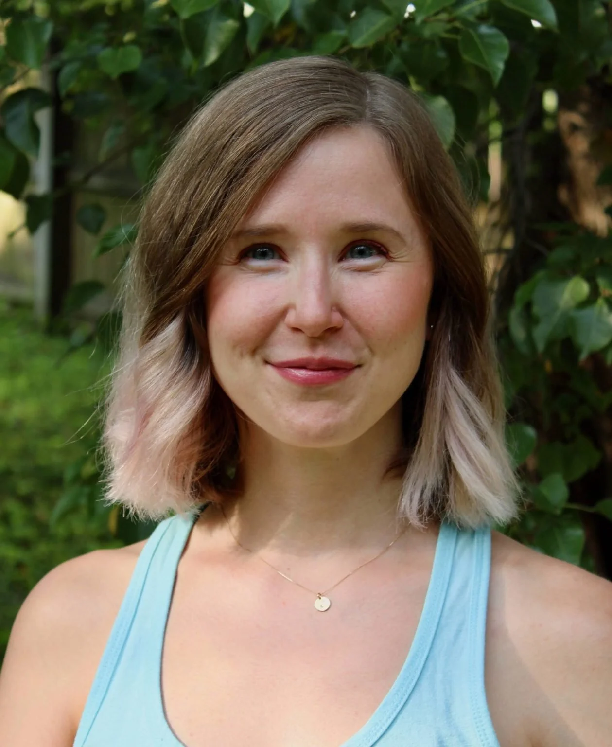 A young woman with shoulder-length light brown and pink hair smiling outdoors, wearing a light blue tank top and a delicate gold necklace with a small circular pendant, with green foliage in the background.