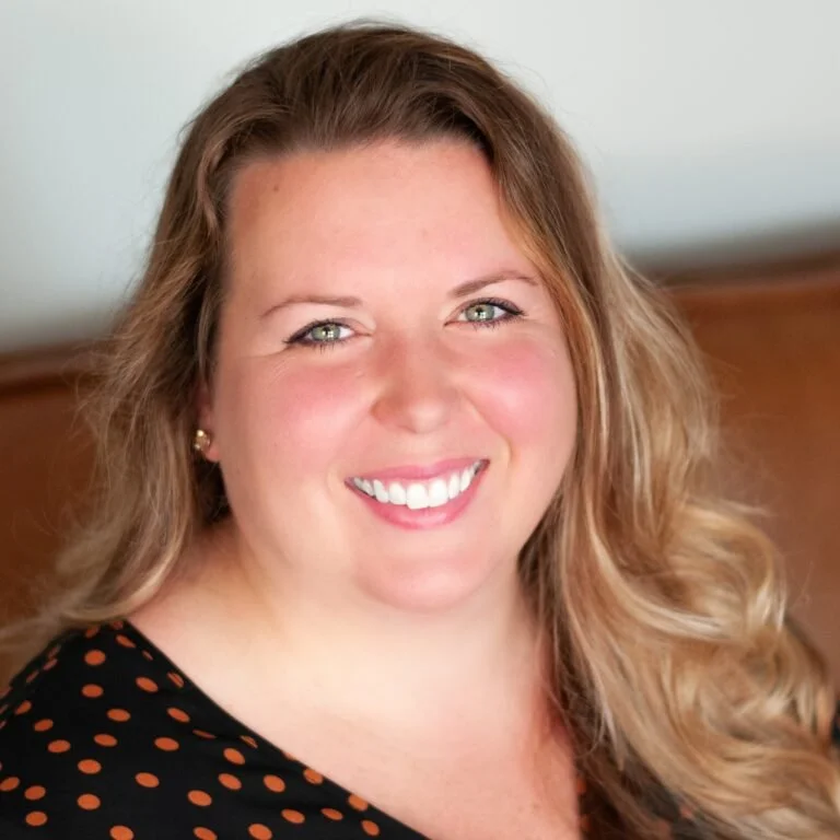 Close-up portrait of a woman with long wavy blonde hair, smiling, wearing a black top with orange polka dots, earrings, against a neutral background.