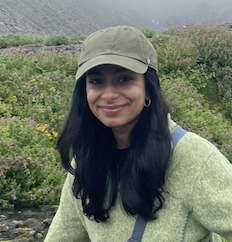 A woman wearing a light-colored cap and a pale green sweater standing outdoors in a lush, flowering landscape.