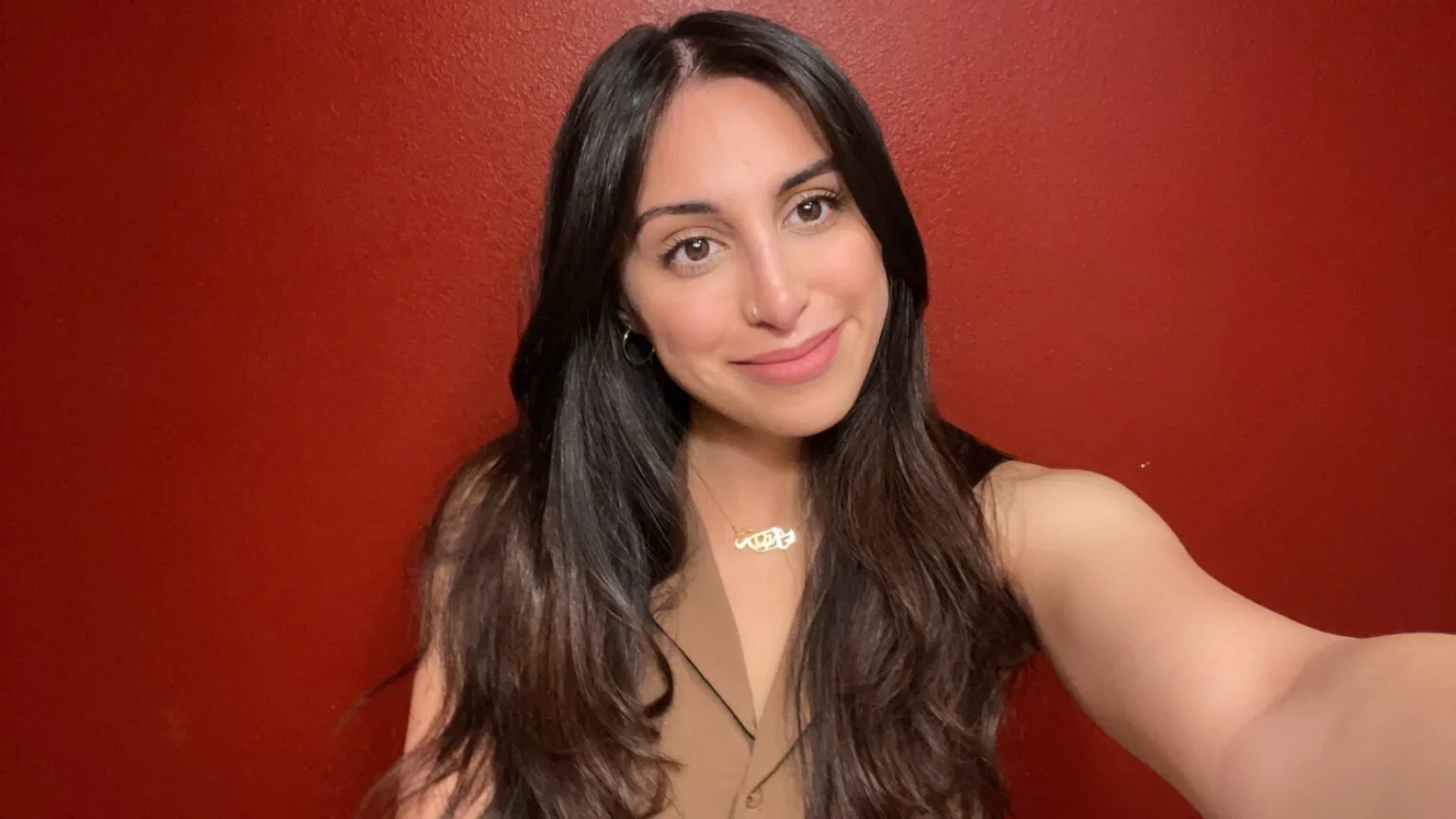 A young woman with long dark hair taking a selfie against a red wall, smiling softly, wearing a necklace and a beige sleeveless top.
