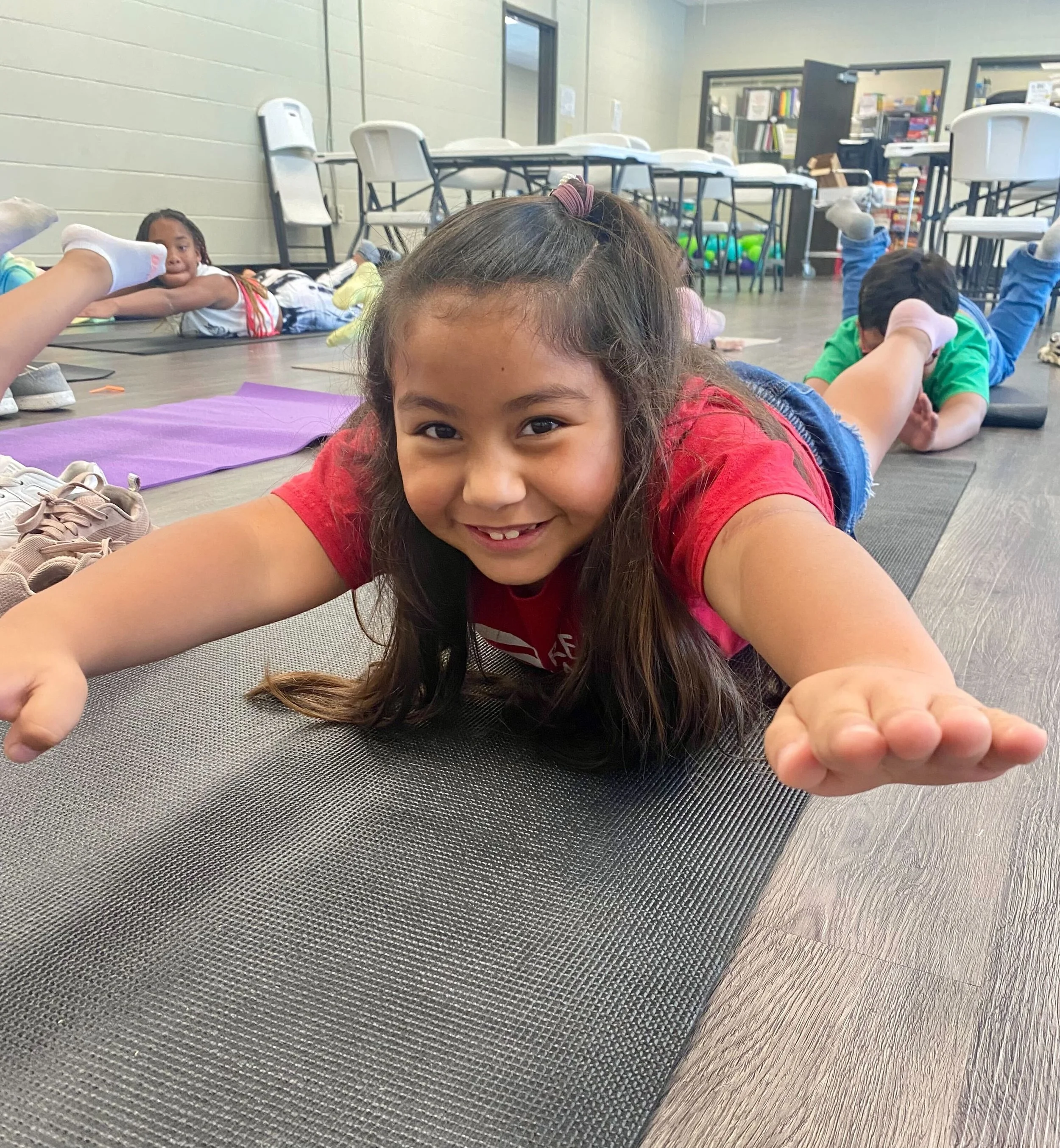 A young girl smiling and lying on her stomach on a yoga mat, participating in a stretching exercise in a classroom or activity room with other children in the background.