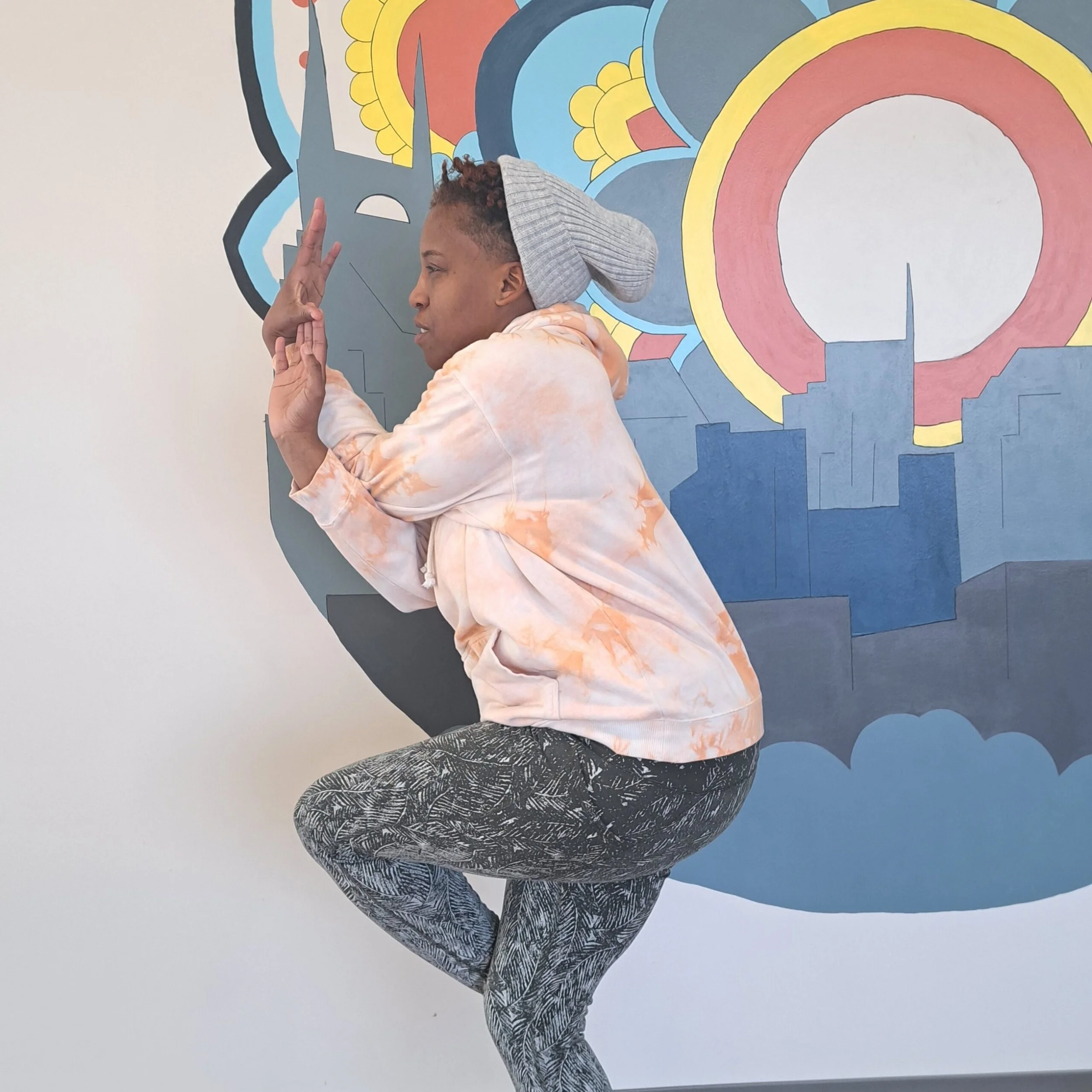 Person doing a yoga pose in front of a colorful mural of a city skyline and a sun or moon.