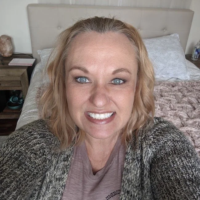 A woman with blonde, wavy hair and blue eyes smiling while taking a selfie in her bedroom. She is wearing a taupe shirt and a patterned jacket. The background shows a bed with white and plush pink blankets, a nightstand with a notebook, a salt lamp, and a water bottle.