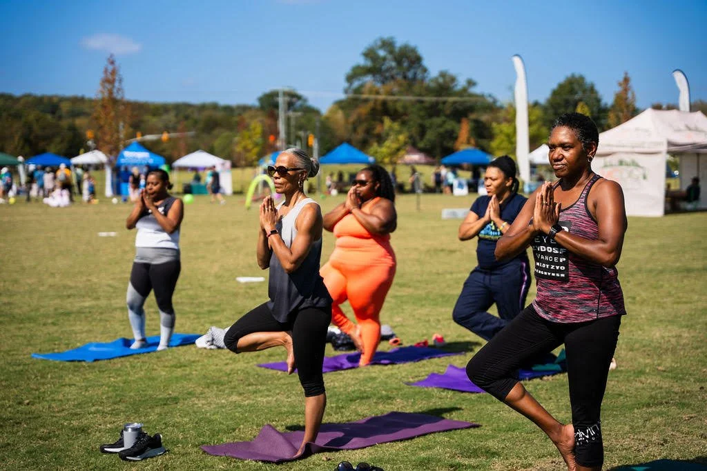 Group of women practicing yoga outdoors on mats during a sunny day with tents and trees in the background.