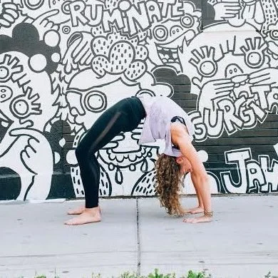 A woman performing a yoga backbend with her hands and feet on the ground, in front of a graffiti wall.