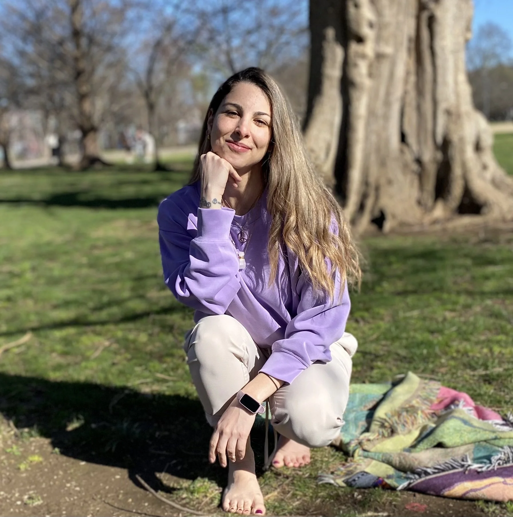 A woman with long blonde hair, wearing a purple hoodie and beige pants, crouches on the grass near a large tree in a park. She is smiling, resting her chin on her hand, and wearing a smartwatch. A colorful blanket is spread out on the grass beside her, and the background shows trees and a blue sky.