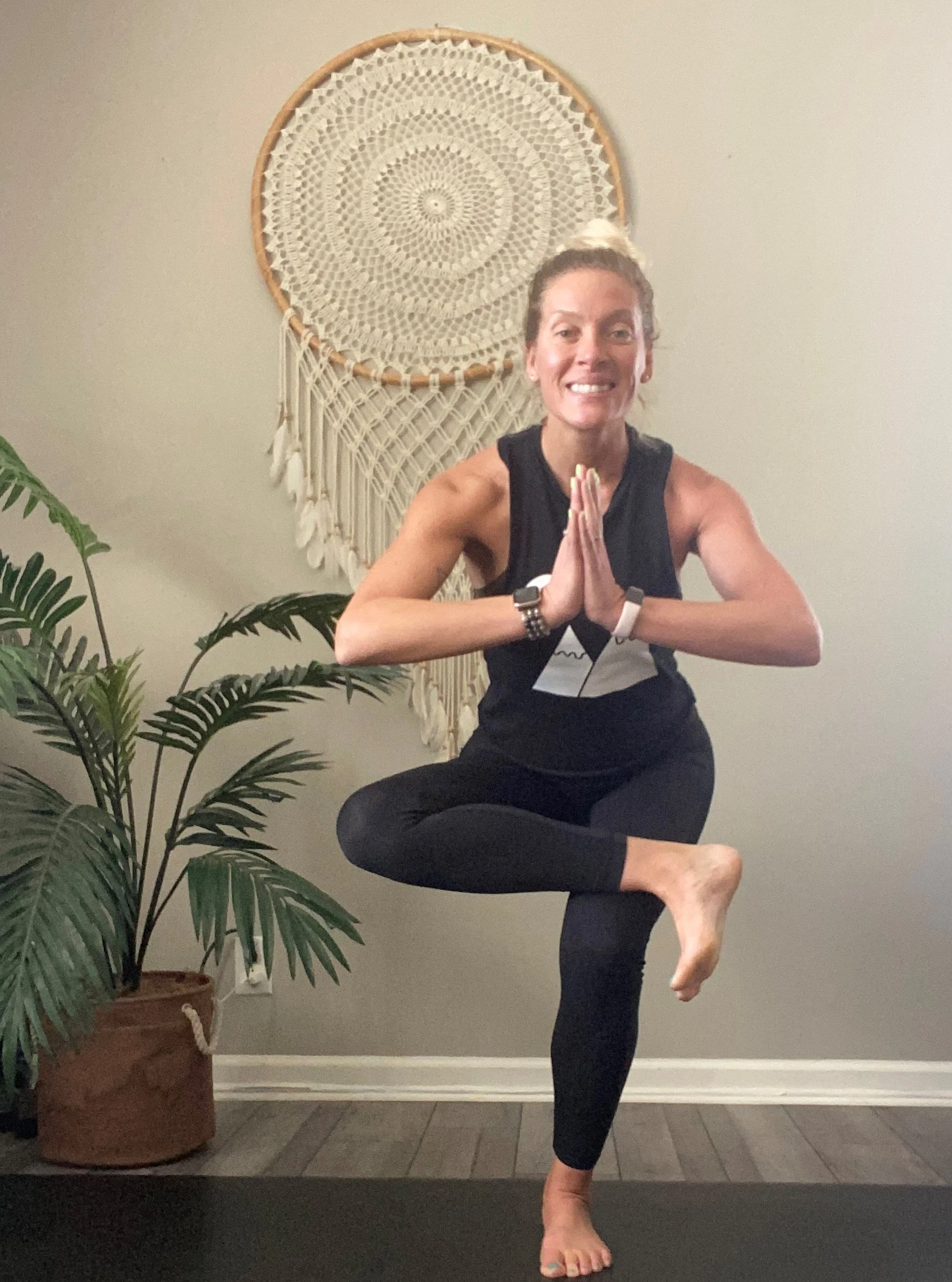 Woman practicing yoga in a yoga pose on a black mat with indoor plants and decorative wall hanging in the background.