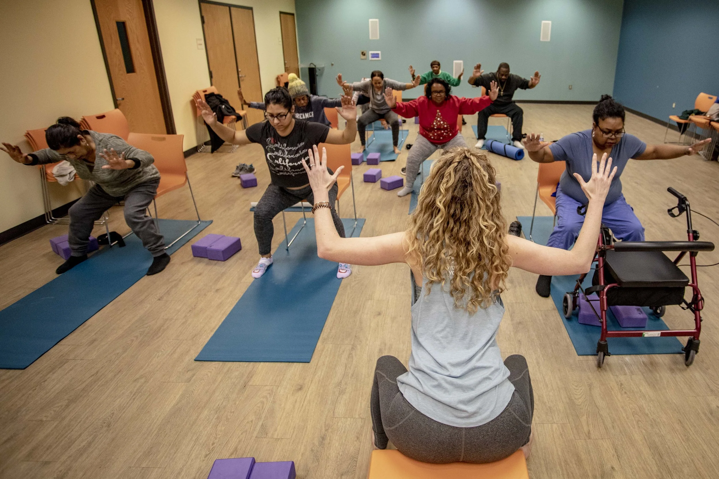 Yoga class in a therapy or senior center with a diverse group of people, including seniors and individuals with mobility aids, following instructor guidance for a seated pose with arms raised.