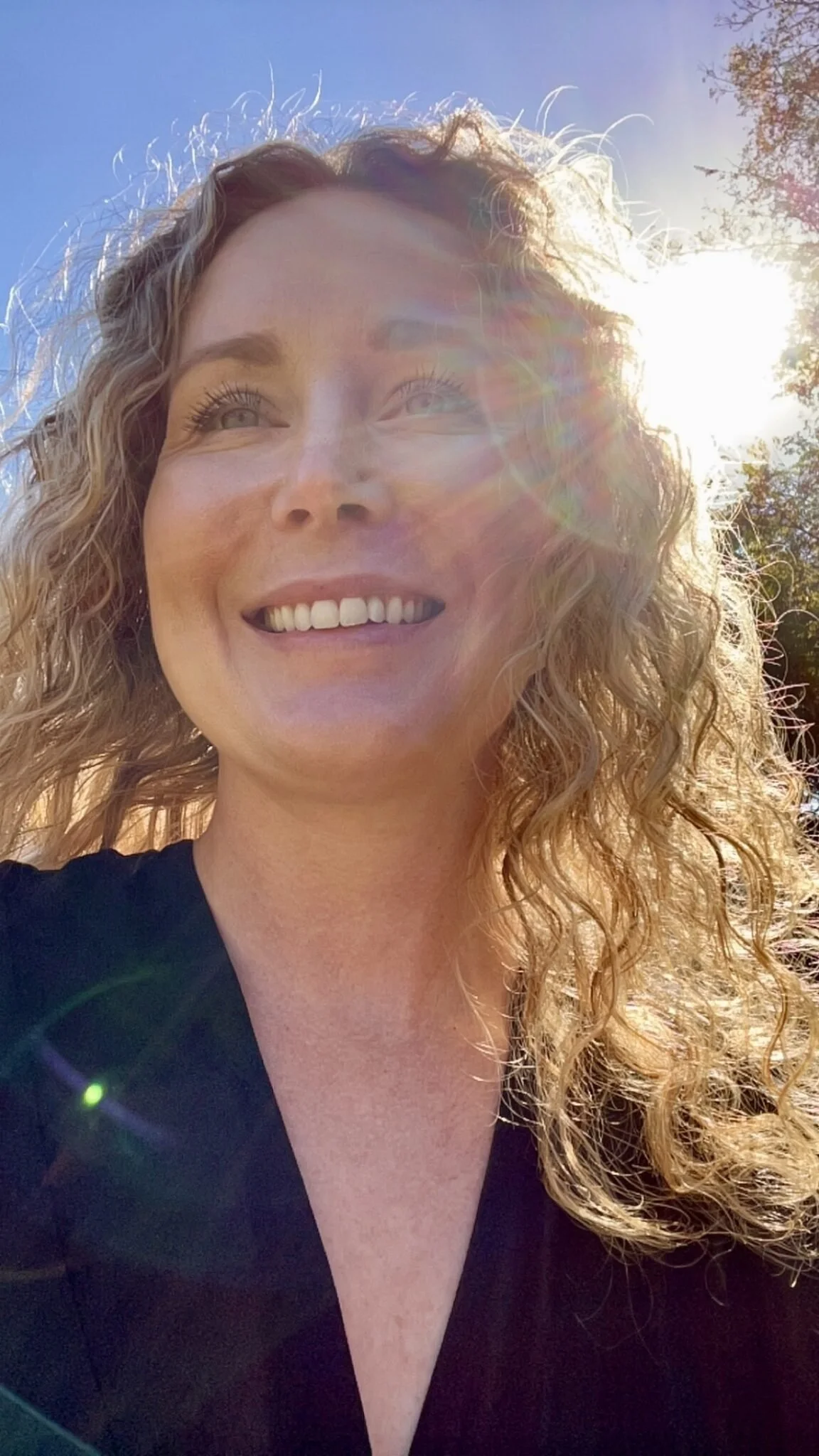A woman with curly blonde hair smiling outdoors with sunlight behind her and a blue sky.