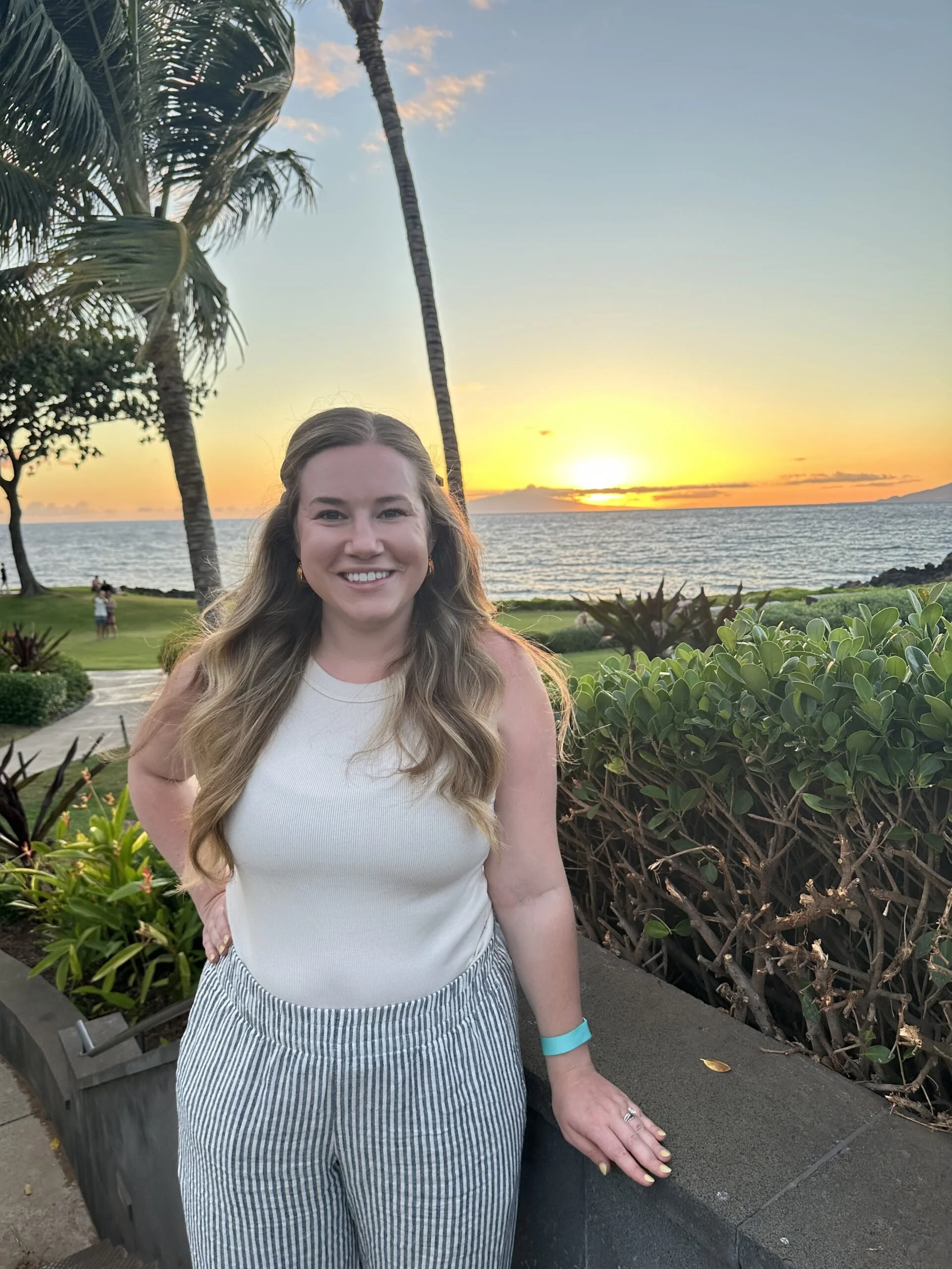 A woman with long wavy hair smiling at the camera while standing outdoors near a garden with bushes and trees, during sunset over the ocean with a clear sky, palm trees, and some people in the background.