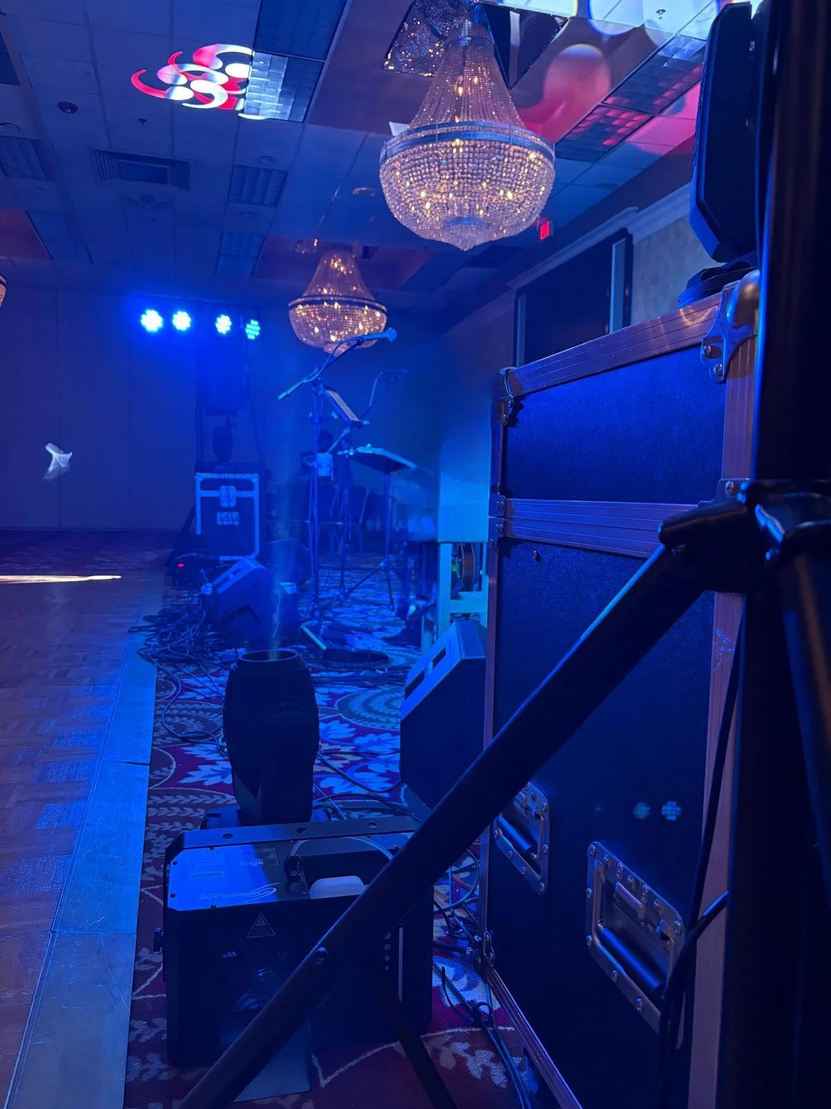 Stage with concert equipment, blue lighting, chandeliers, and a microphone on a stand in a dimly lit room.