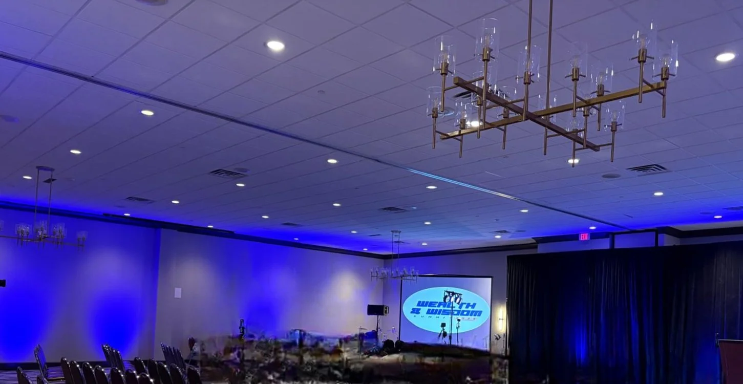 Empty conference room with dim blue lighting, decorated with chandeliers, with a large screen displaying a logo for 'A WEALTH WED' and chairs lined up along one side.