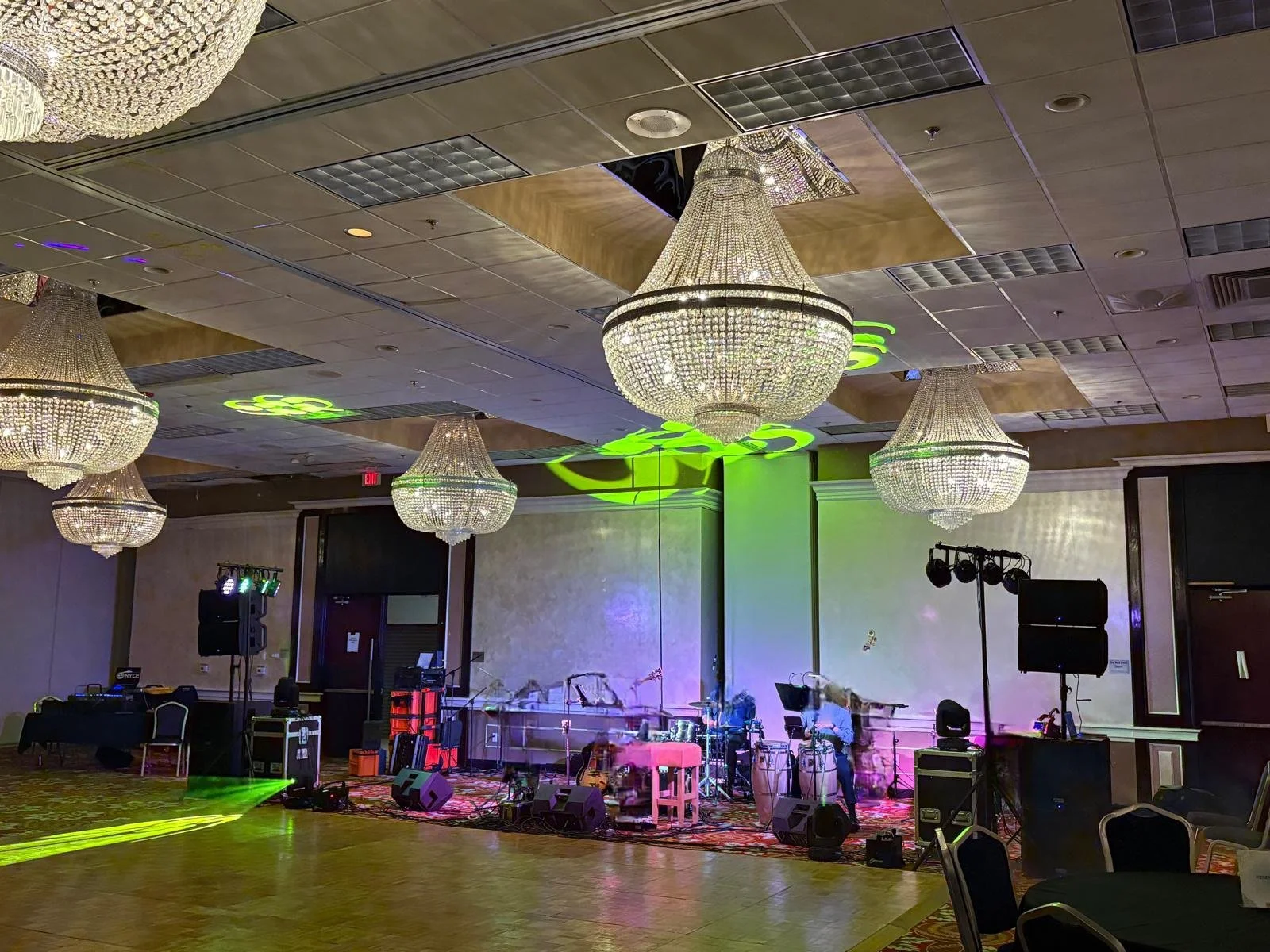 A banquet hall with four large chandeliers hanging from a decorated ceiling. The stage is set with musical instruments, speakers, and lighting equipment, ready for a performance or event.